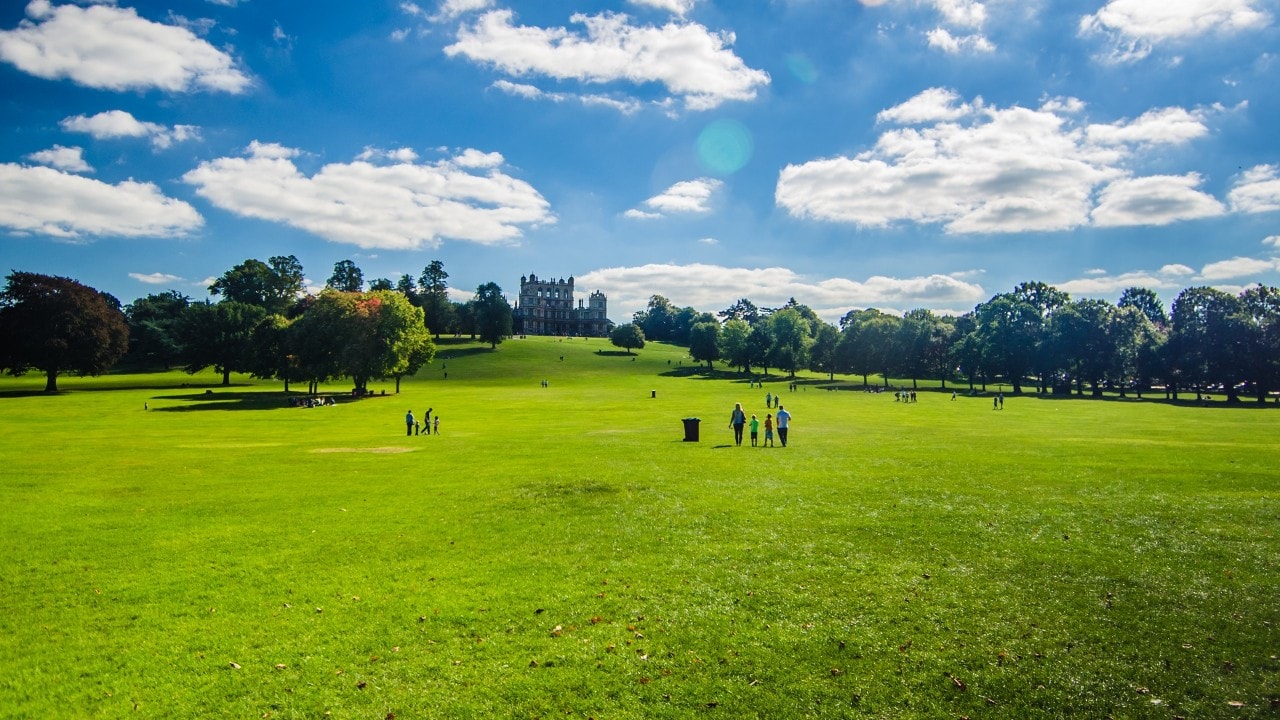 People walking at Wollaton Hall Park in Nottingham on a sunny day with green field, amazing blue sky and white clouds.